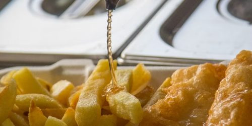 Malt vinegar being poured over freshly cooked chips beside golden battered fish at The Fishermen’s Chippy