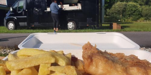Fish and chips served in a takeaway box with The Fishermen’s Chippy food truck in the background