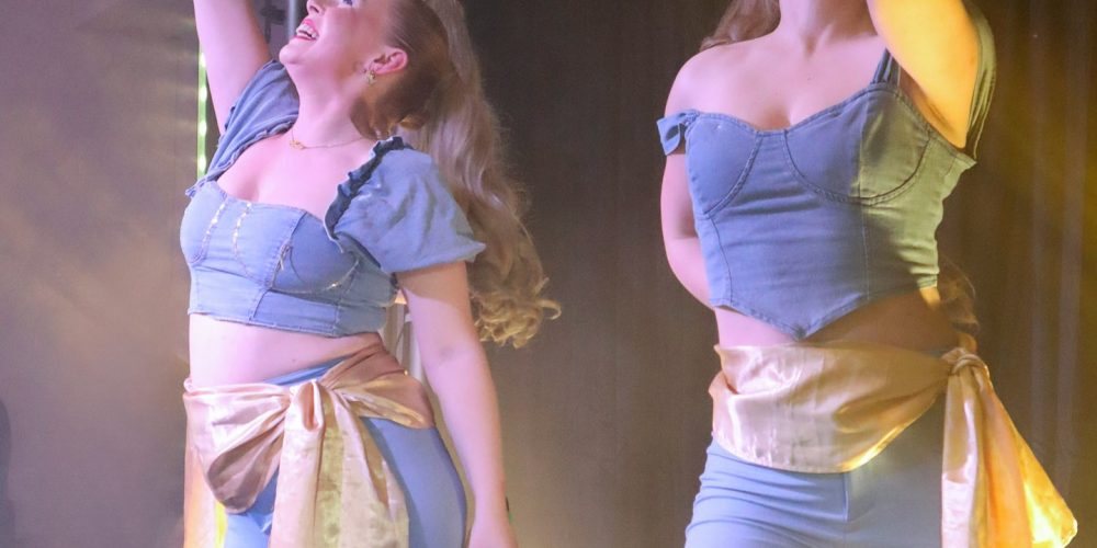 Two female singers performing live on stage with microphones during an evening event, under colourful stage lighting.